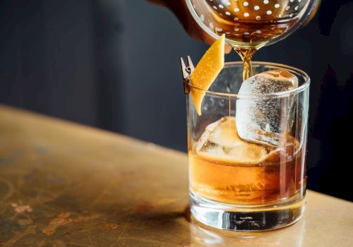 A cocktail is being poured into a glass with a large ice cube and a citrus twist garnish on a bar counter.