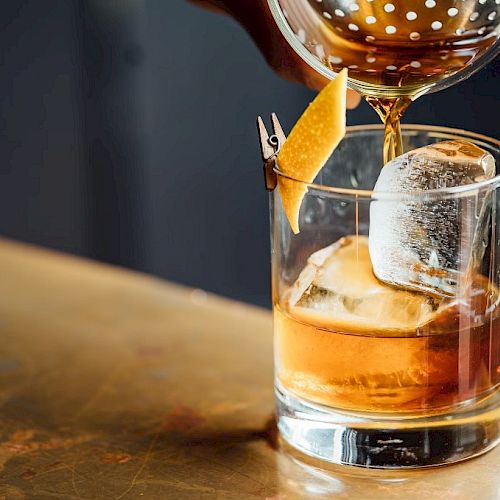 A cocktail is being poured into a glass with a large ice cube and a citrus twist garnish on a bar counter.