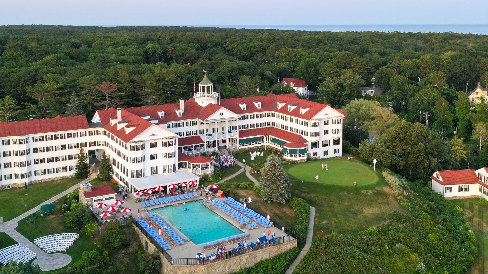 A large building with red roofs and white walls, surrounded by trees, featuring a pool area and seating arrangements outside.