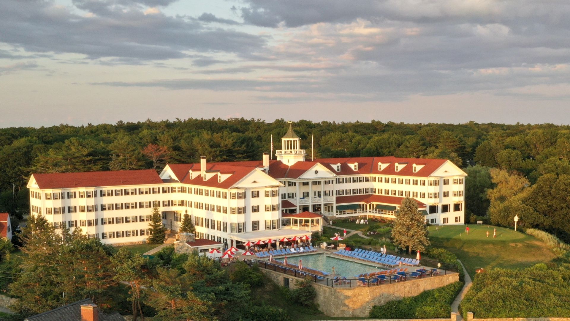 A large, historic-looking building with a red roof, surrounded by trees. There's a swimming pool and a patio with chairs in the foreground.
