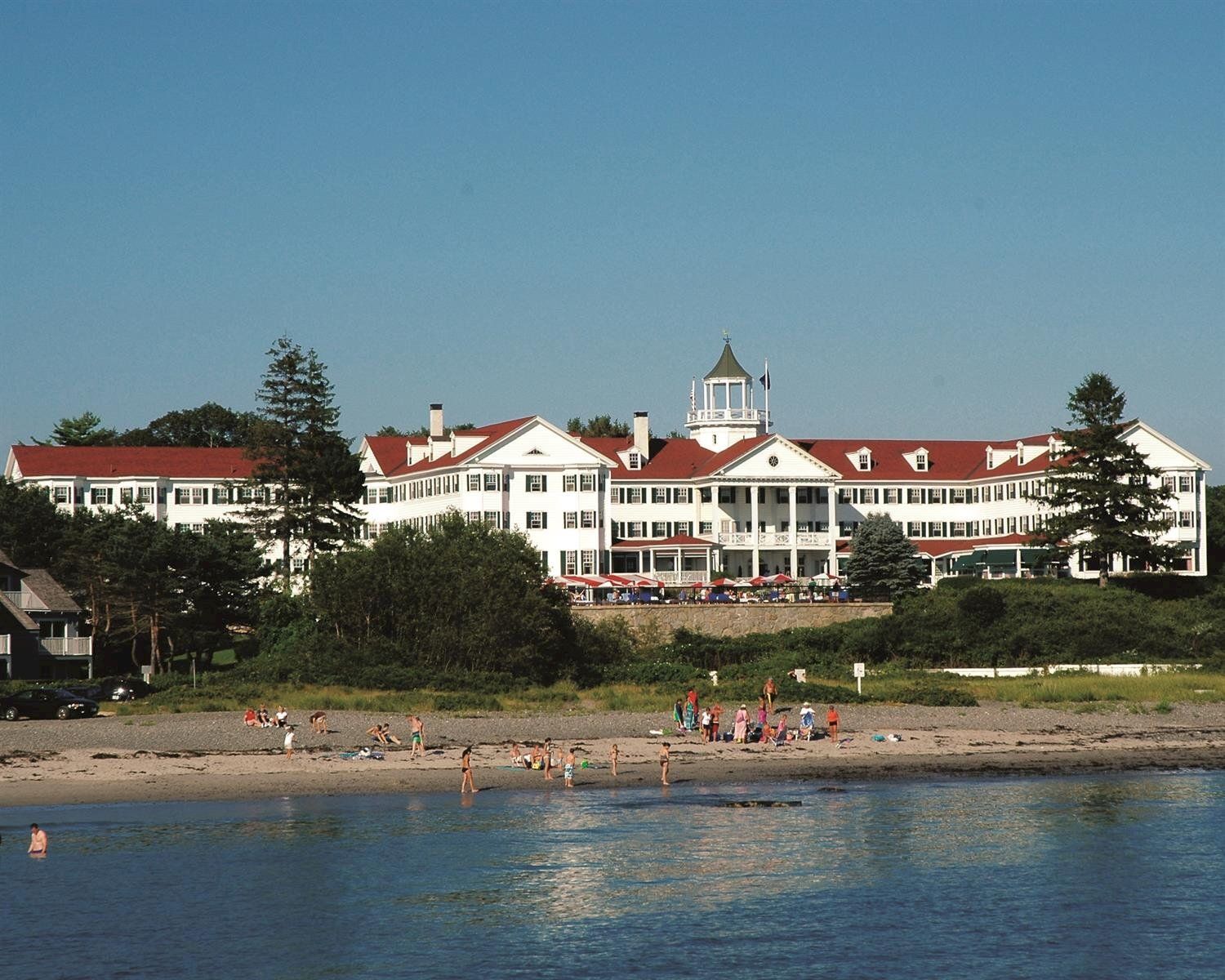 The image shows a large building with a red roof near a beach, with people enjoying the shoreline and trees in the background.