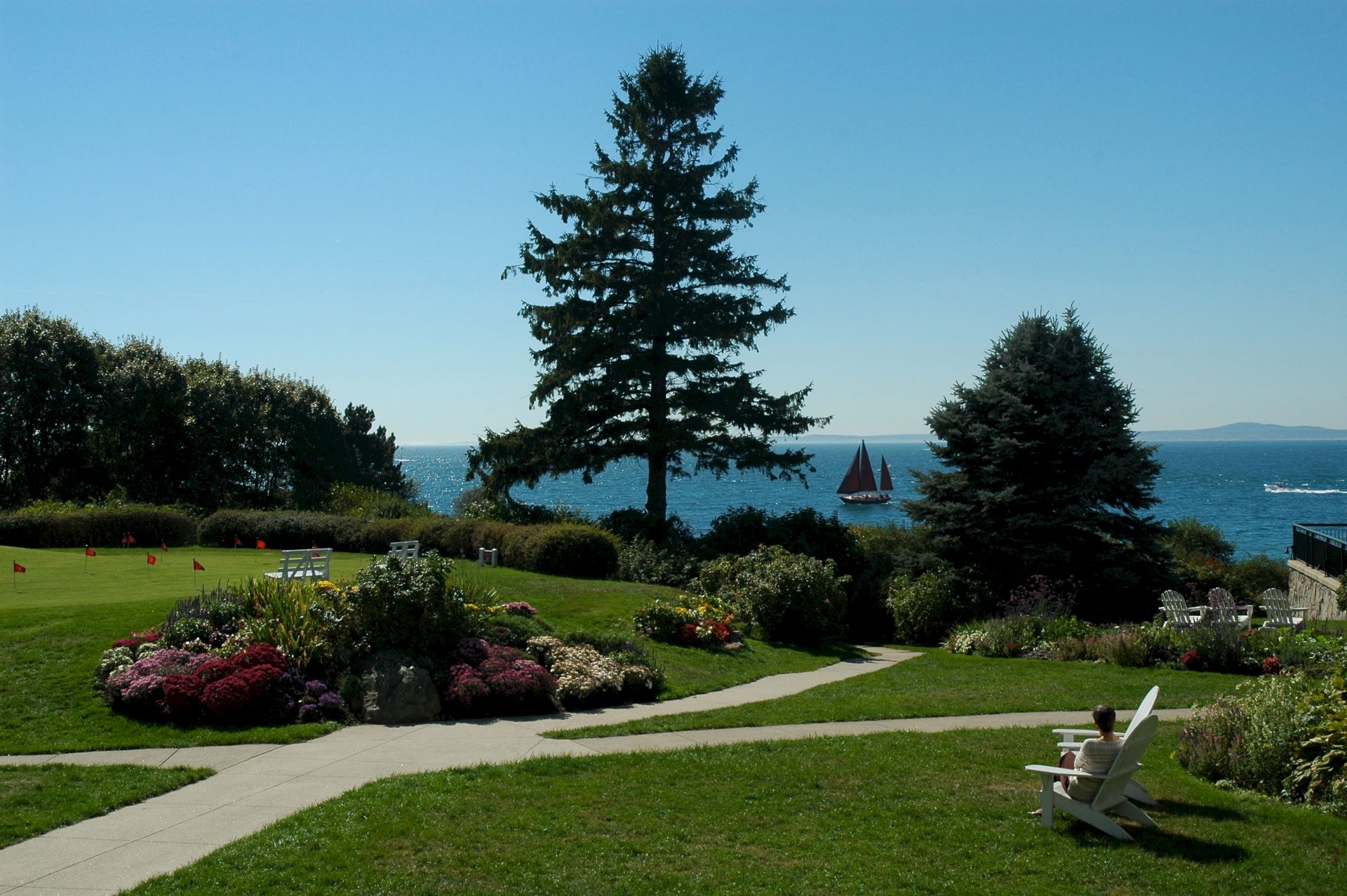 A scenic view of a garden with pathways, trees, and a lake in the background with a sailboat. Two white chairs face the water.