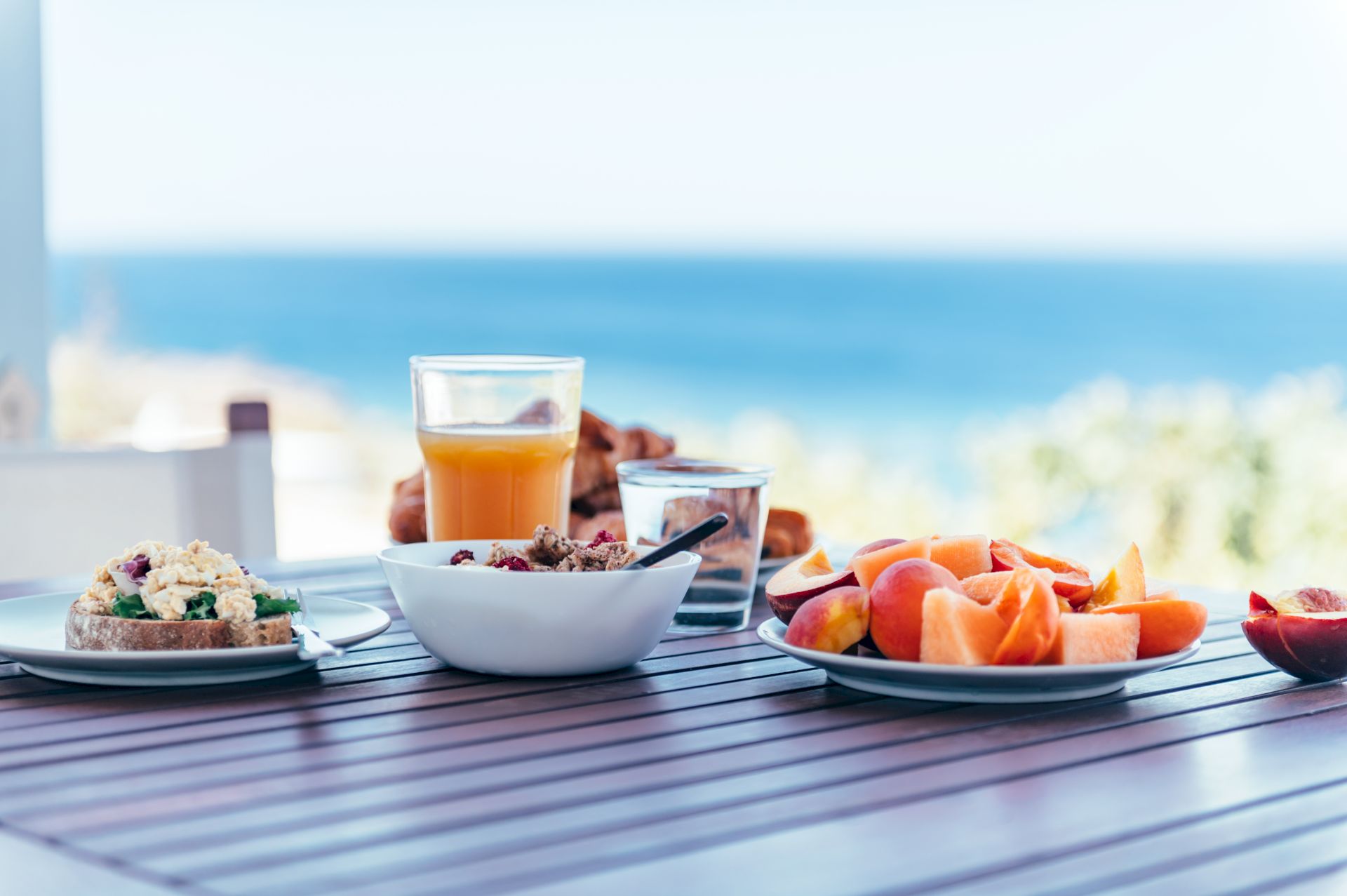Breakfast on a wooden table overlooking the sea: toast, fruit, yogurt, and juice create a fresh and inviting scene.