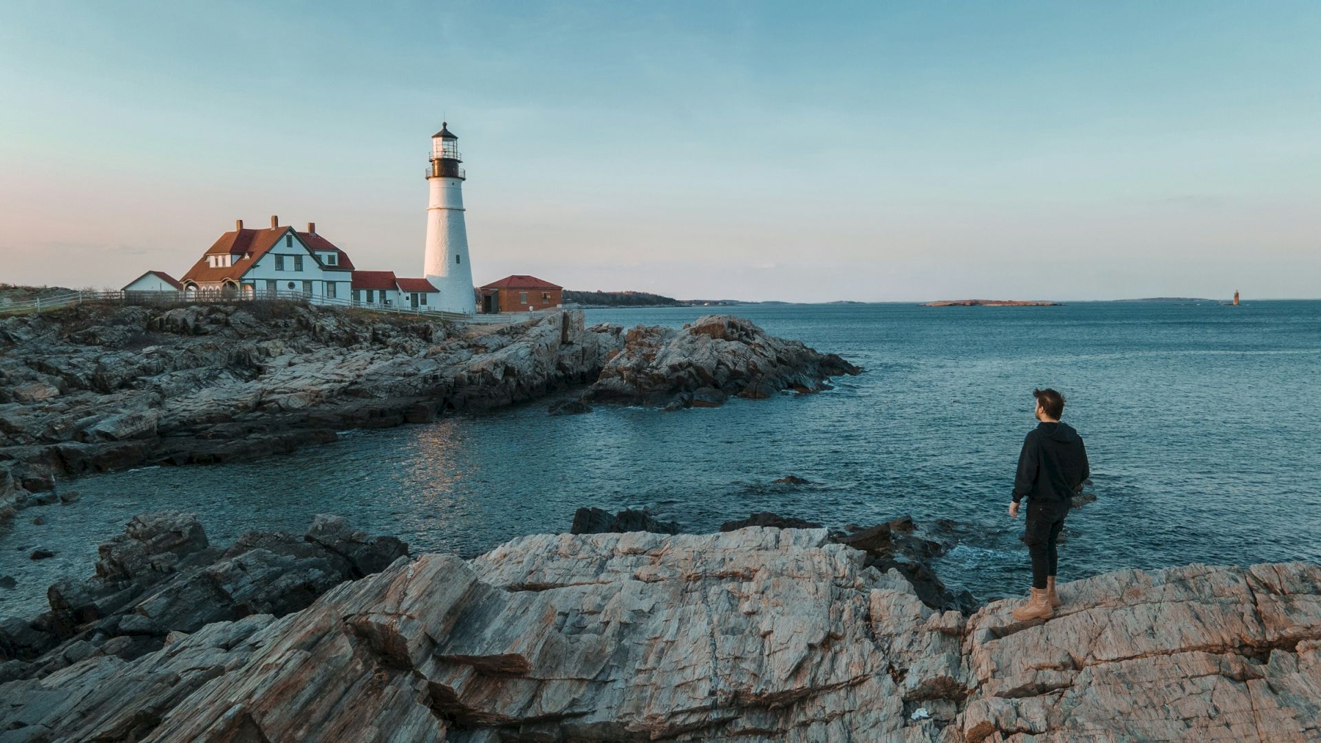 A person stands on rocky cliffs, gazing at a lighthouse by the sea with adjacent buildings under a clear sky.