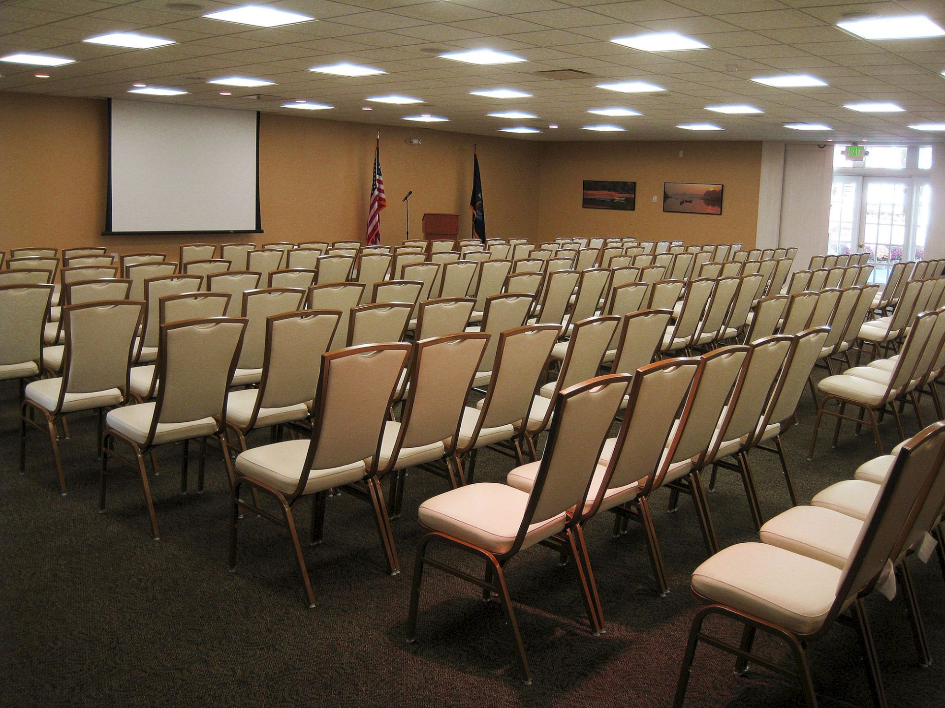The image shows an empty conference room with rows of chairs facing a screen and flags, ready for an event or meeting.