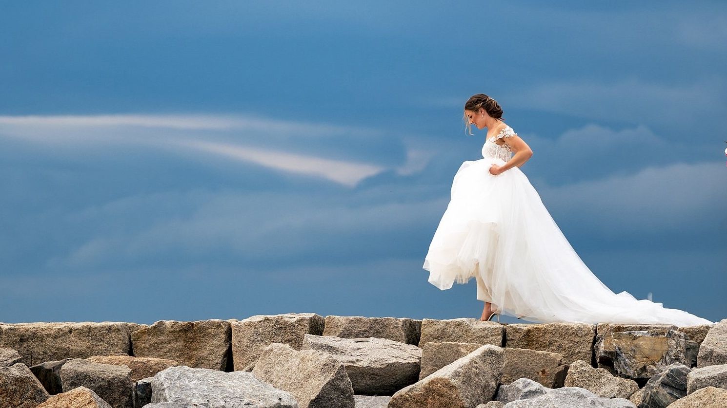 A couple stands on rocks under a stormy sky; she's in a wedding dress, he's in a suit holding a bouquet.