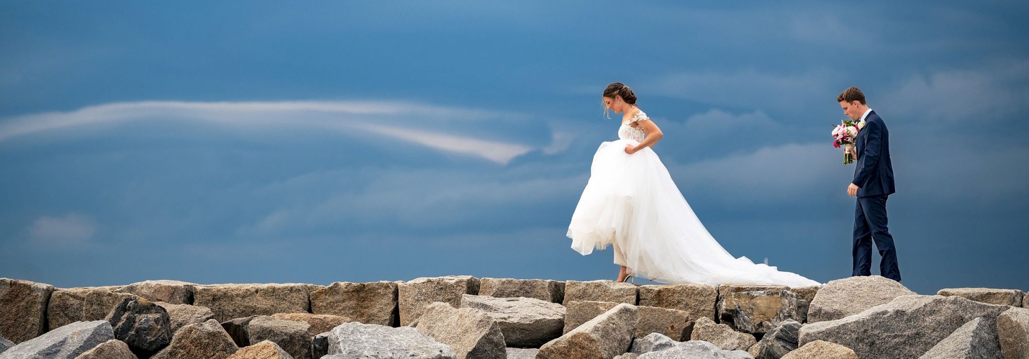 A couple stands on rocks under a stormy sky; she's in a wedding dress, he's in a suit holding a bouquet.