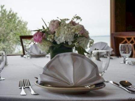 A formal table setting with a napkin, plates, glasses, and cutlery, and a floral centerpiece in the background.