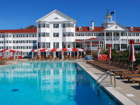 A large hotel with red roofs and white facade is seen behind a swimming pool and rows of lounge chairs under red and white umbrellas.