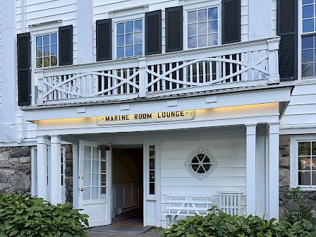 White building entrance with dark shutters and a sign reading &ldquo;Marine Room Lounge&rdquo; above an open door.