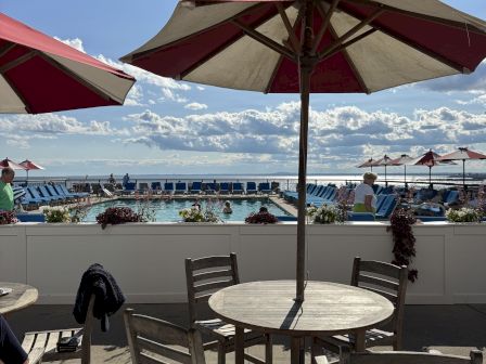 Seaside pool deck with blue lounge chairs, red umbrellas, and an ocean view under sunny clouds, with a wooden round table in the foreground.
