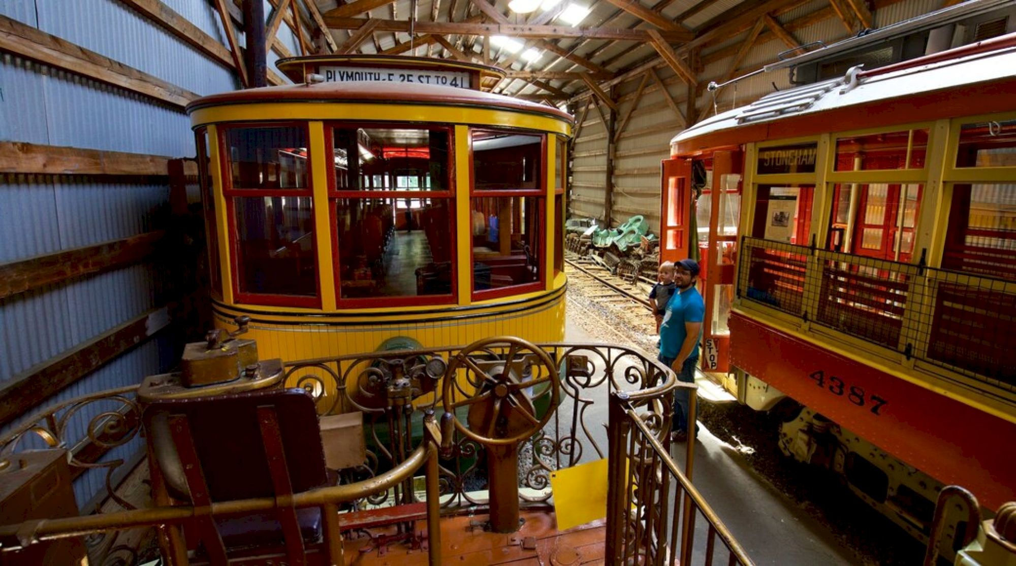 The image shows two vintage streetcars in a barn-like structure; one is yellow, the other red, with a person standing nearby.