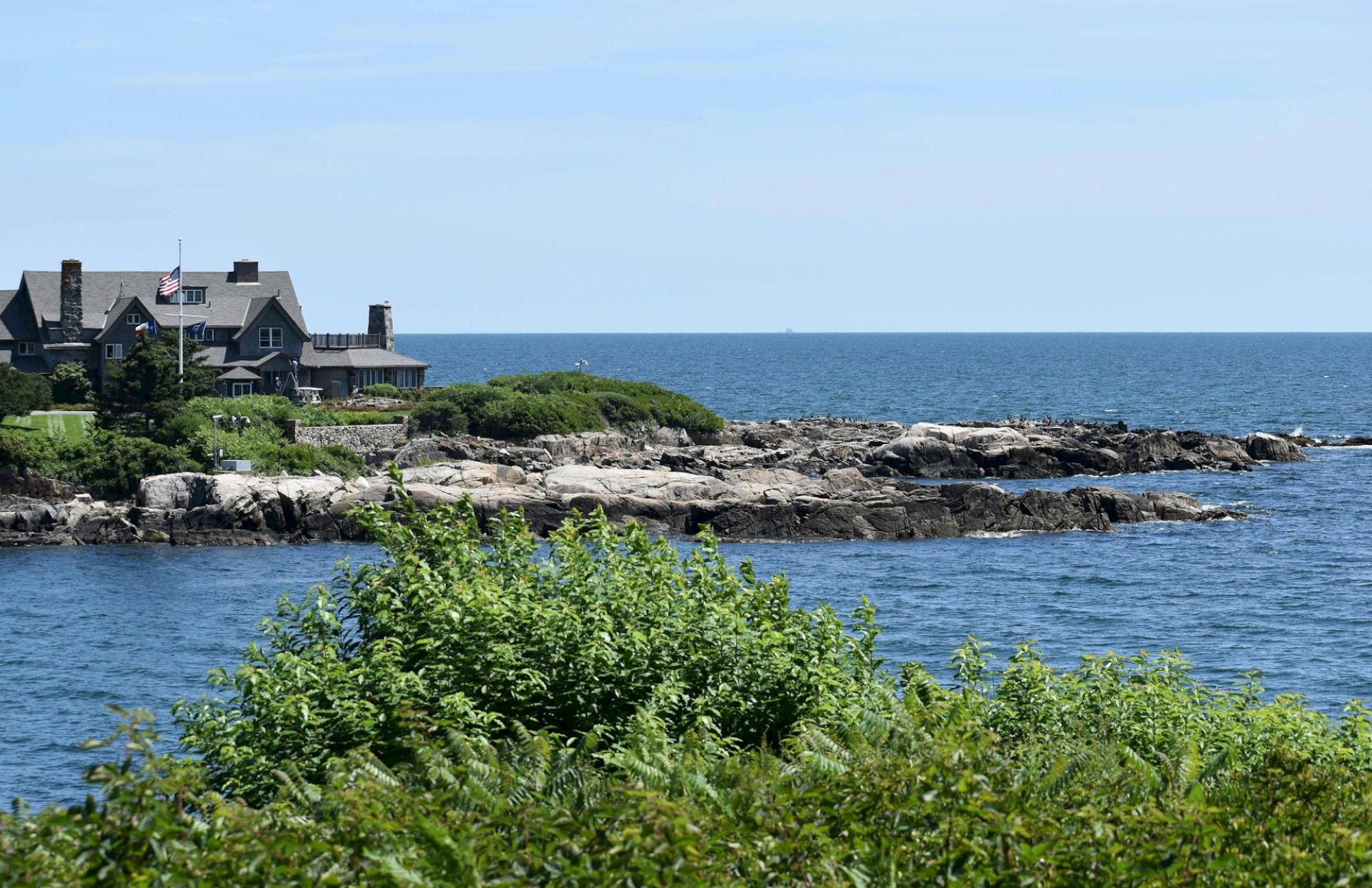 A coastal house with an American flag overlooks rocky shores and the ocean, surrounded by greenery under a clear blue sky.