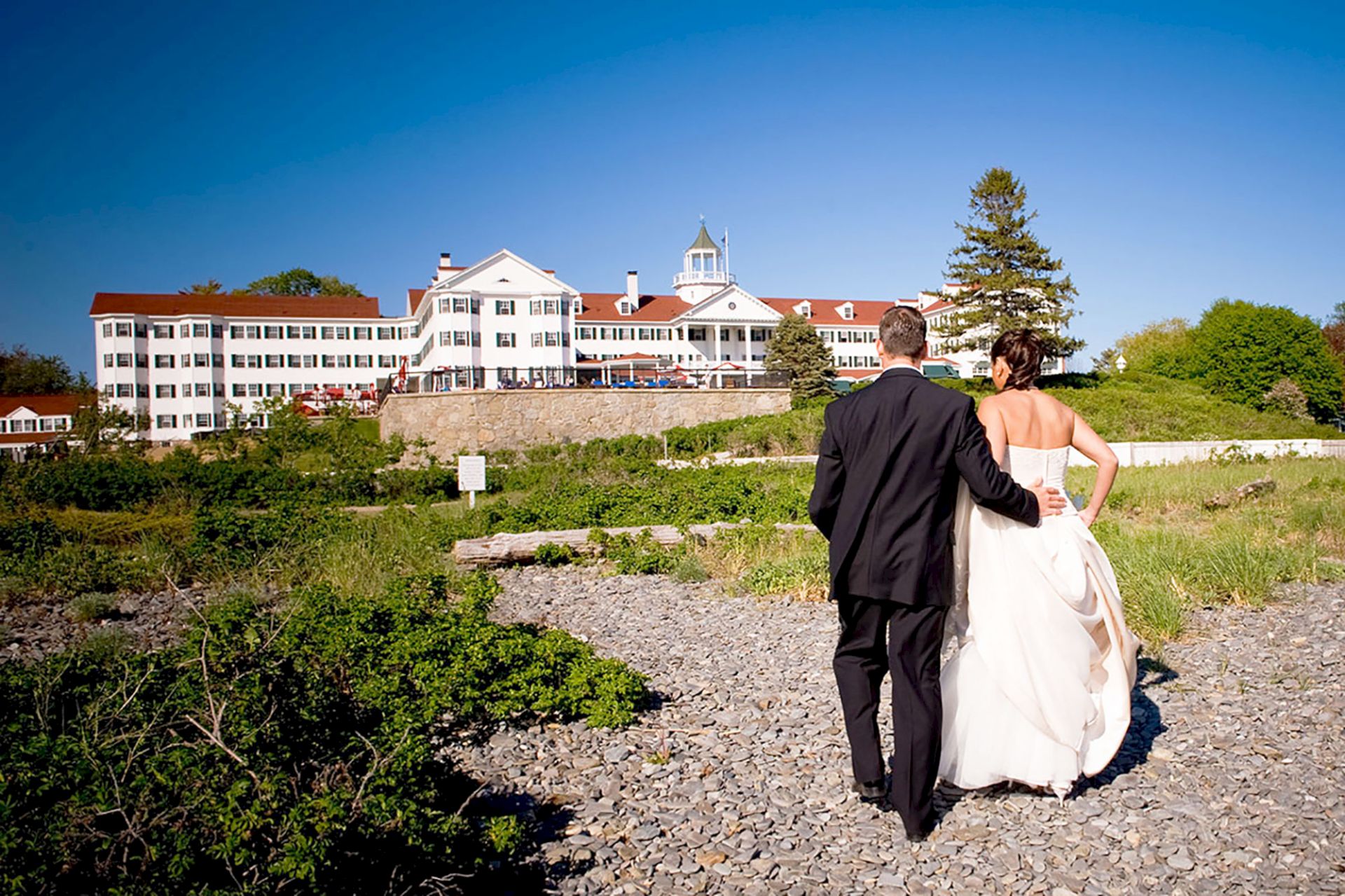 A couple in wedding attire walks on a rocky path toward a large white building on a sunny day.
