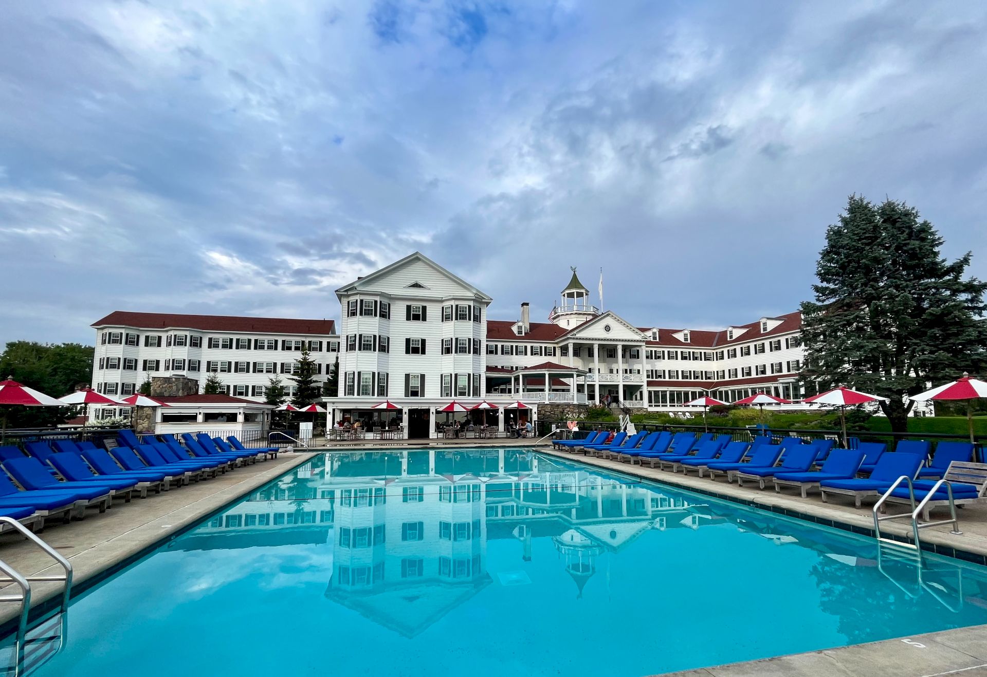 A grand hotel with white siding and red roof, featuring a central dome, behind a pool lined with blue lounge chairs and red umbrellas.