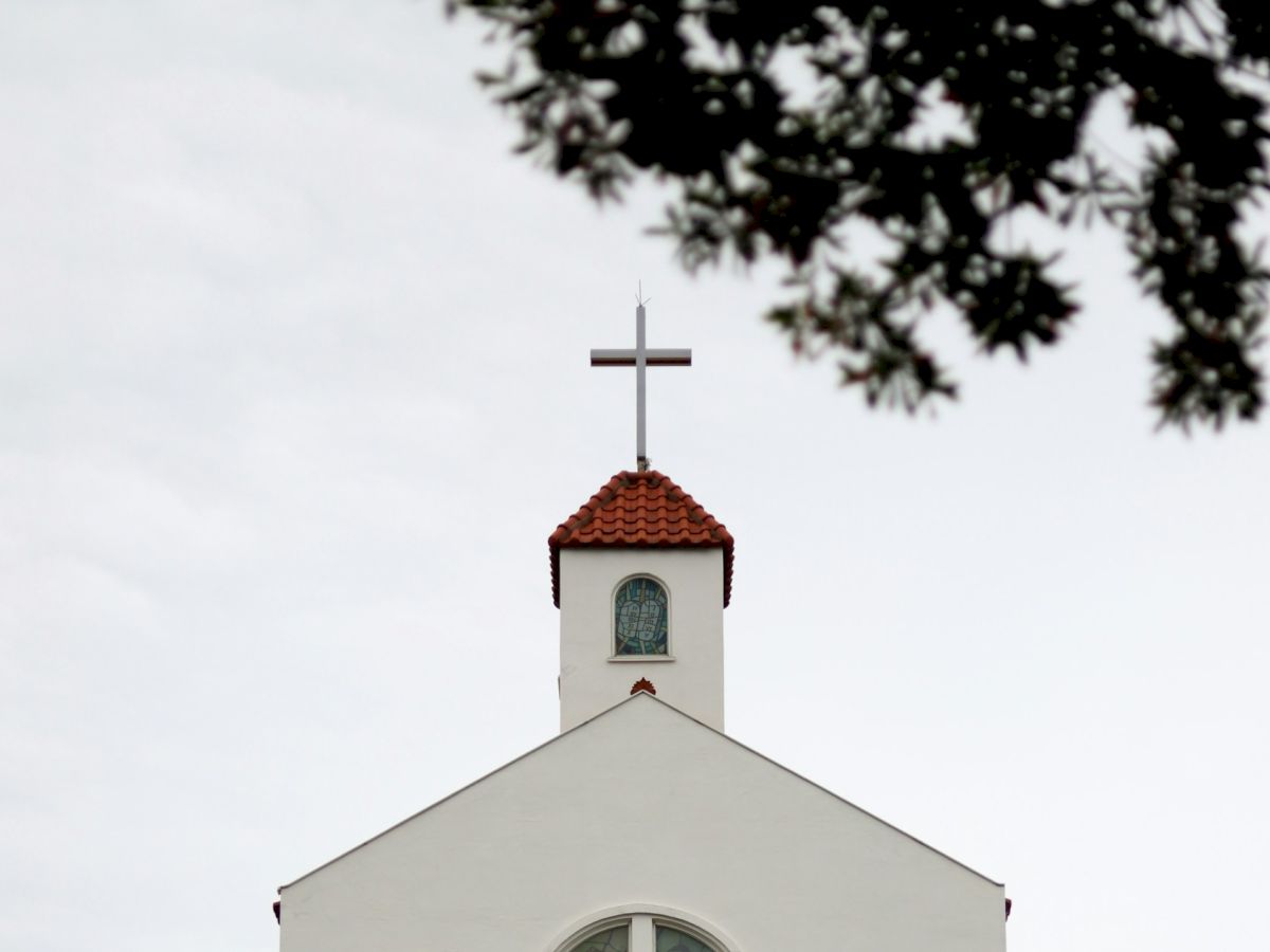 The image shows the top of a church with a cross, a small tower with a red-tiled roof, against a cloudy sky and some tree branches.