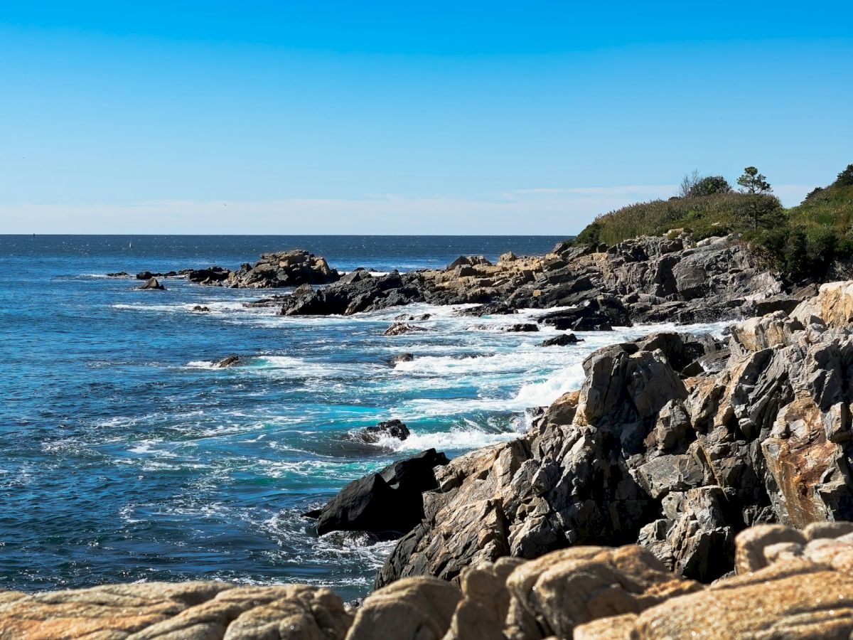 A rocky coastline with blue ocean waves under a clear sky, lined with greenery and distant rocky outcrops.