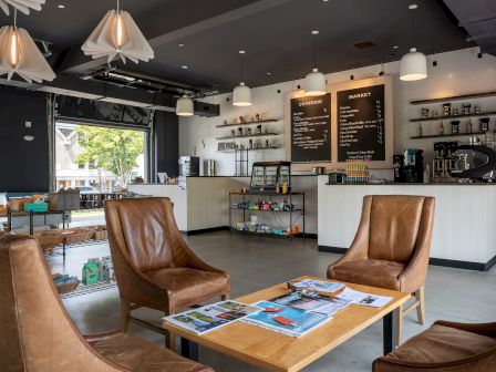 A cozy cafe with leather chairs, wooden tables, a counter with menus, and shelves displaying items. It has pendant lighting and an open layout.