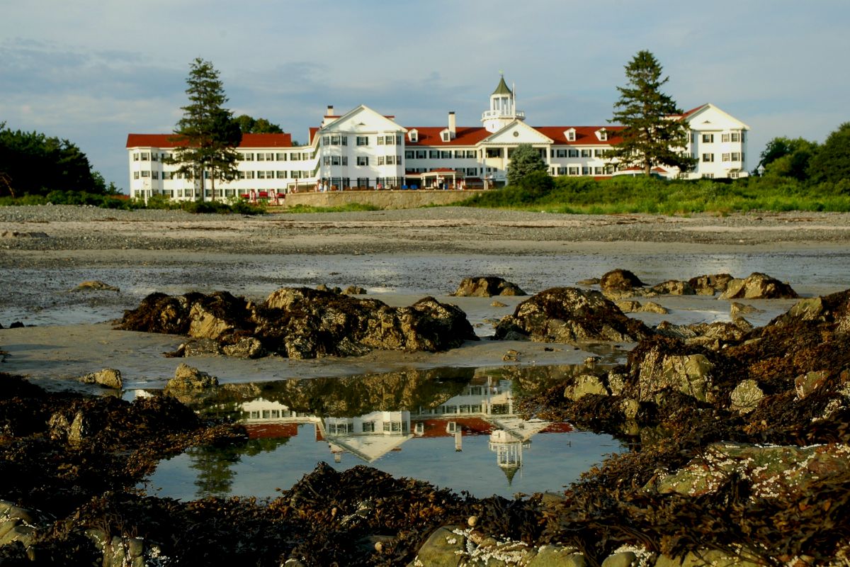 A large hotel with a red roof is reflected in a tide pool on a rocky shore, surrounded by greenery.