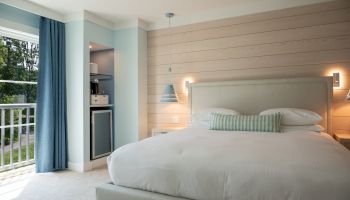 A bright hotel room with a large bed, light wood panel wall, blue pendant lights, a balcony with blue curtains, and a small seating nook by a mini-fridge.