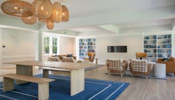 A bright, modern living space with a large wooden table, woven pendant lights, comfy seating, built-in bookshelves, and a blue rug.