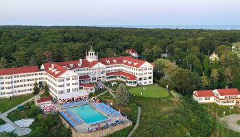 The image shows a large building with red roofs, surrounded by greenery, featuring a pool area with lounge chairs and a scenic landscape.
