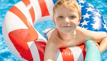 A child is in a pool, using a patriotic-themed inflatable ring and holding a pool noodle, smiling at the camera.