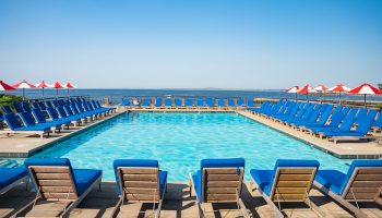 A bright hotel pool area with blue lounge chairs around a rectangular pool and red umbrellas near the horizon, ocean in the background.