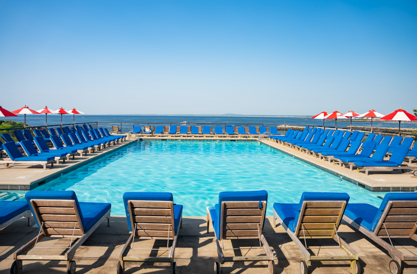 A bright hotel pool area with blue lounge chairs around a rectangular pool and red umbrellas near the horizon, ocean in the background.