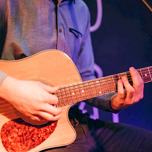 A person is playing an acoustic guitar, focusing on strumming and fretwork, under stage lighting.