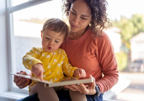 A person holds a child on their lap by a window, both looking at a book, with the child pointing at it, creating a cozy reading moment.