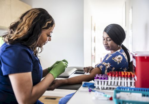 A healthcare professional draws blood from a patient in a clinic, with medical supplies visible on the desk next to them.
