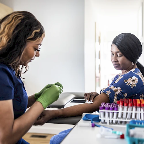 A healthcare professional draws blood from a patient in a clinic, with medical supplies visible on the desk next to them.