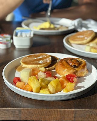 Plates with pancakes, a pastry, and fruit, set on a table with cutlery, and a person in the background.