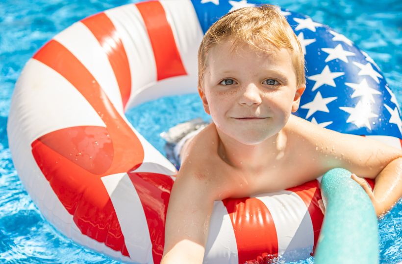 A child is in a pool, holding onto a noodle, and using a red, white, and blue inflatable ring with stars and stripes.
