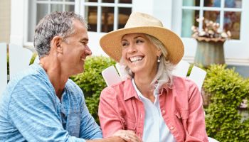 A smiling couple sits together outdoors. The woman wears a straw hat and the man is in a blue shirt, both appearing happy and relaxed.