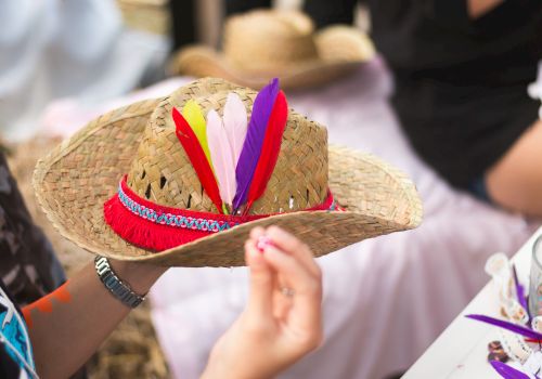 A straw hat with colorful feathers and trim is held by a person at a craft table.