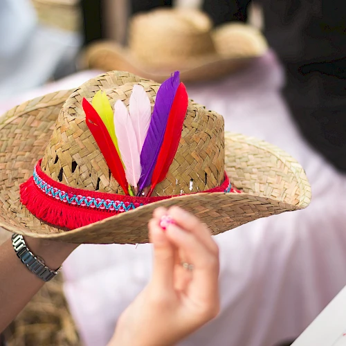 A straw hat with colorful feathers and trim is held by a person at a craft table.