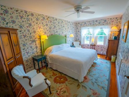 A cozy bedroom with floral wallpaper, a bed with white linens, a wooden wardrobe, and two chairs by a window with ample natural light.