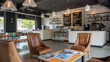 A cozy café interior with brown chairs, a wooden table, and a menu on the wall; the counter has coffee machines and shelves display items.