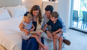A family of four, sitting on a bed, is engaged in a book together in a bright room with a patio door in the background.