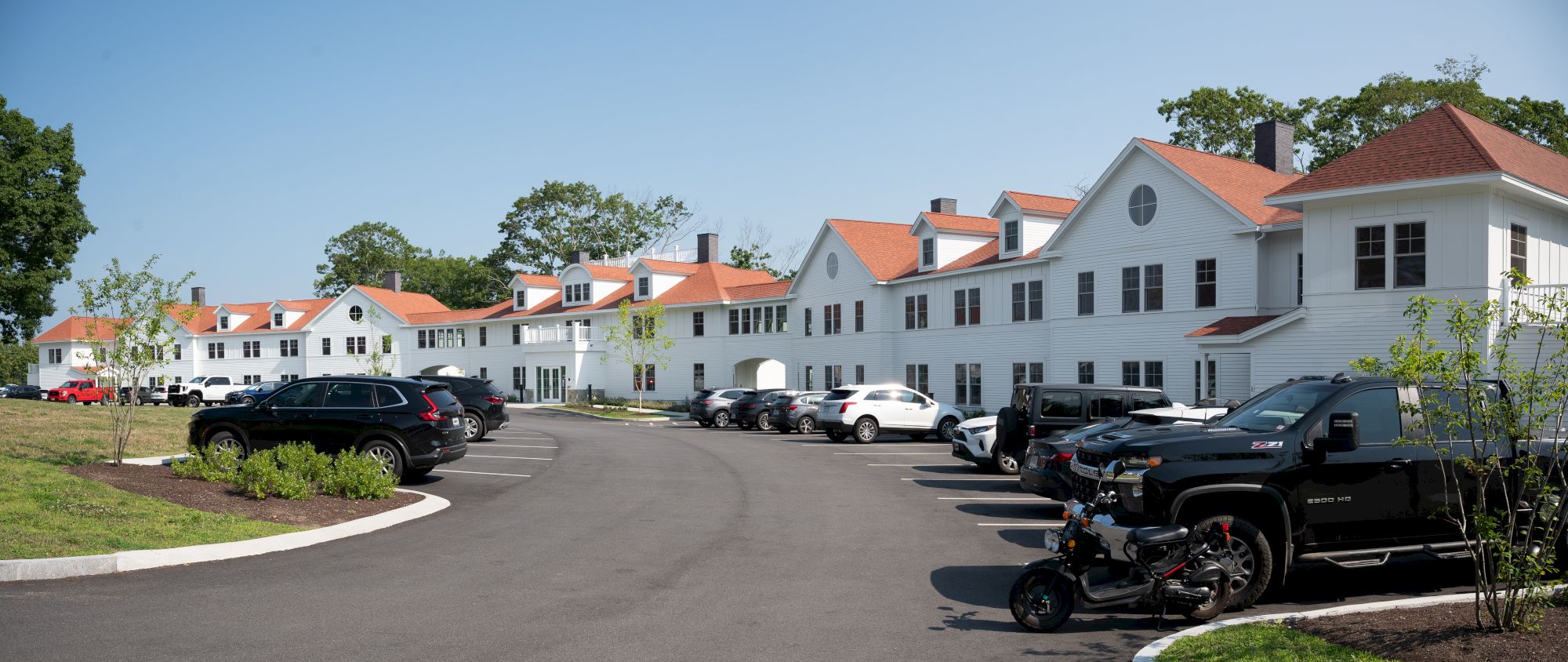 The image shows a row of white buildings with red roofs, parked cars, and trees under a clear blue sky.