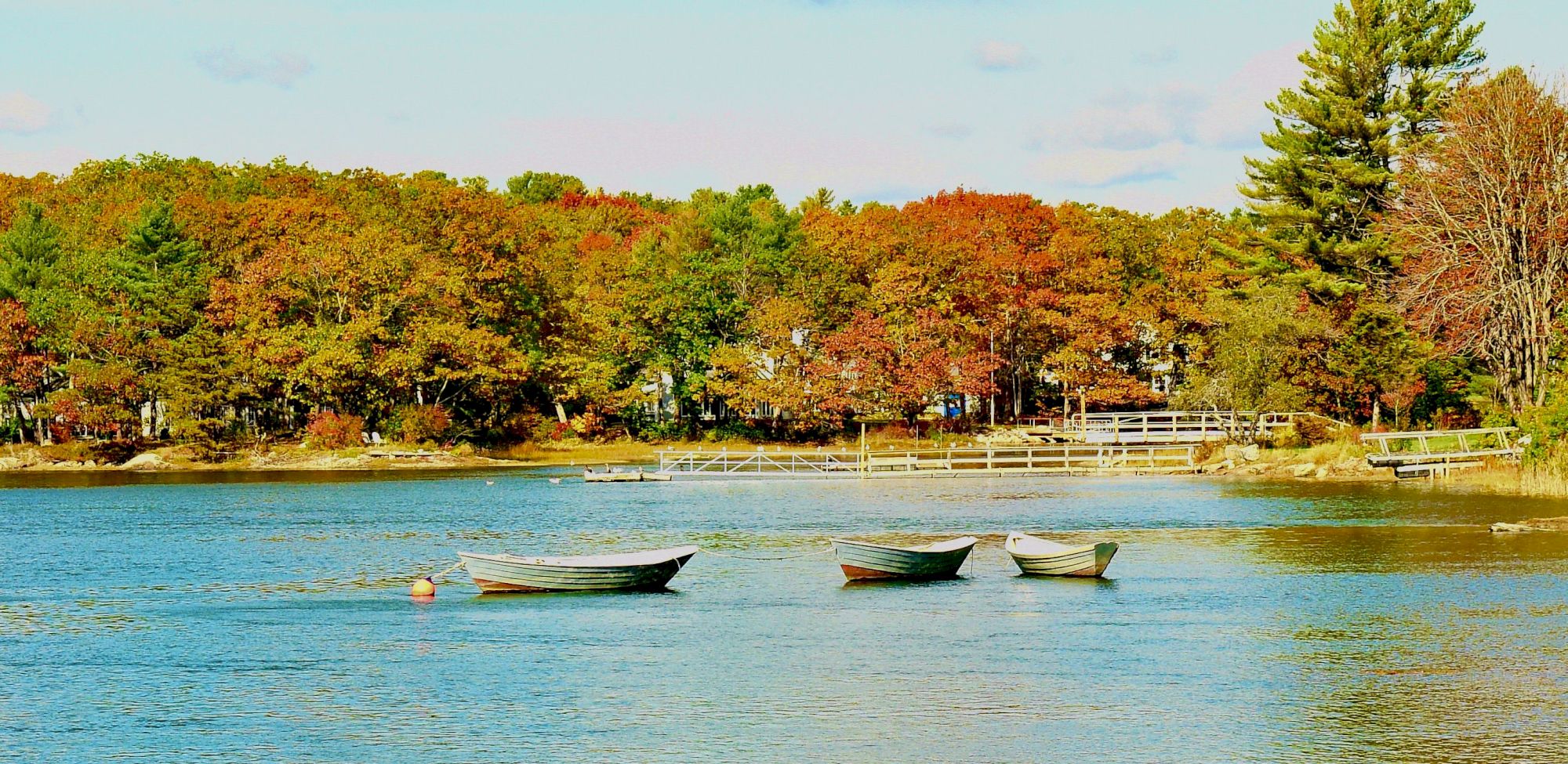 A serene lake scene with three boats floating and a backdrop of colorful autumn trees and a small bridge in the distance.