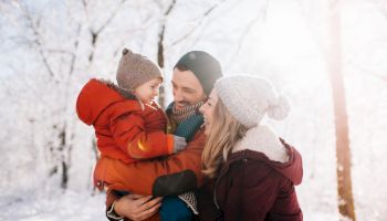 A smiling family of three in a snowy forest: a mother, father, and toddler in warm coats and hats, enjoying a sunny winter day together.