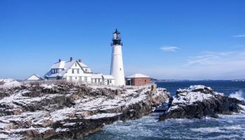 A white lighthouse and adjacent keeper buildings sit on rocky coast, with icy foreground, blue sky, and the ocean beyond, calm and bright.