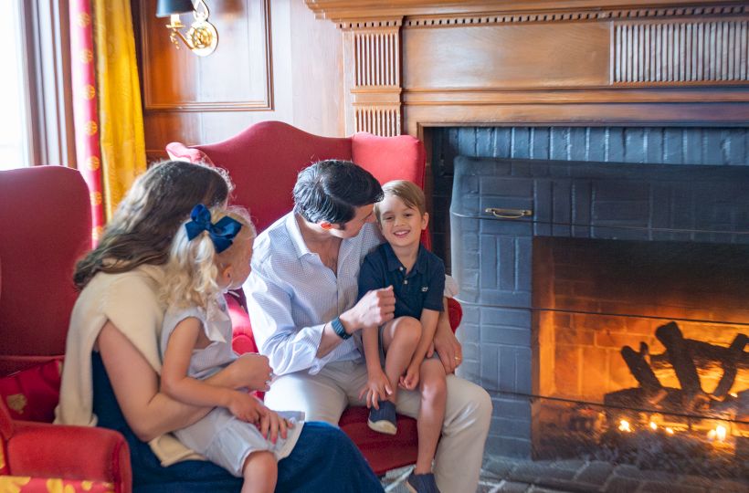 A family sitting by a cozy fireplace in a warmly decorated living room with a painting and vases on the mantel.