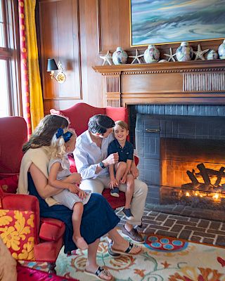 A family sits together by a lit fireplace, with children on their laps, in a cozy, decorated living room.