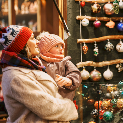 A woman and child in winter clothing admire colorful ornaments at a festive market display, surrounded by holiday decorations.