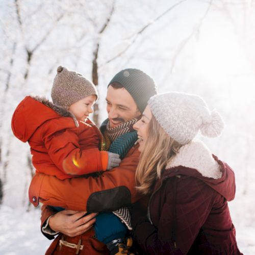 A family of three in a snowy forest: a smiling mom and dad hold their child in warm winter coats and knit hats, sunlight glows softly.