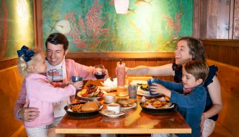 A family of four enjoying a meal at a restaurant, smiling and raising glasses, with plates of food on the table.